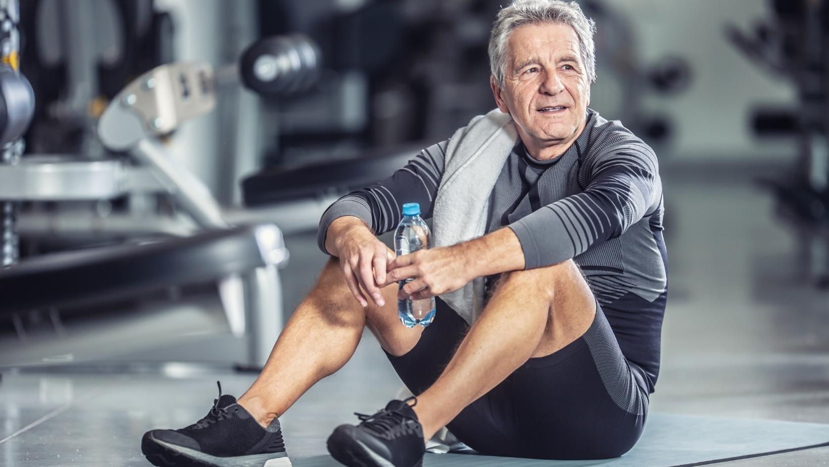 Ein älterer Mann sitzt im Fitnessstudio am Boden und hält eine Wasserflasche in der Hand